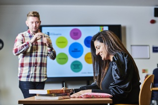 A student working at a desk with a stack of textbooks, while another student stands in front of an interactive whiteboard in the background