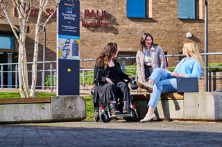 Photograph of three students outside the Lord Swraj Paul building, two seated on a bench and the third using a wheelchair.