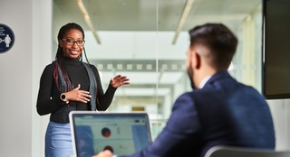 One student standing and speaking to another student who is sat on a desk on his laptop.