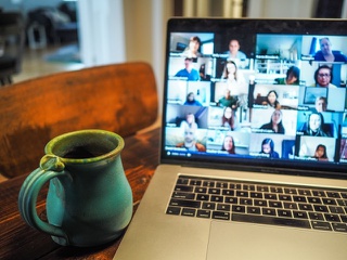 A cup of coffee sits by an open laptop, the laptop screen displays the faces of a number of participants in a call.