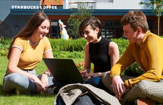 Image of students sitting on grass and conversing; a laptop computer is being used by one student