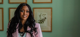 Amanda Nkomo wearing a pink blazer, standing in front of a green wall and an arrangement of framed art prints.