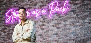 Nate Ethan Watson wearing a white jacket, standing in front of a brick wall where a pink neon sign hangs