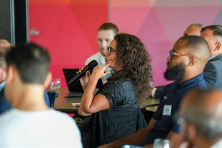 A woman with long, dark curly hair and glasses is seated at a business event. She is asking a question whilst holding a microphone