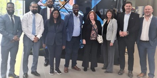 A group of visitors to the University stand in front of a banner