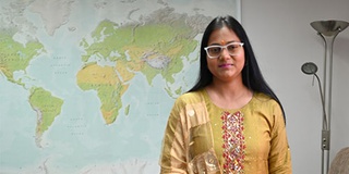 International student, student Vanjyotsna wearing a traditional yellow Indian dress standing in the Chaplaincy in front of a world map
