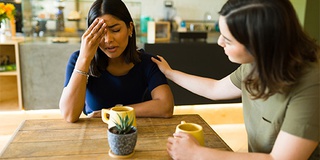 Two women in sitting in a coffee shop, one comforts the other who appears distressed