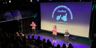 A photograph taken in a theatre at Deaffest. The photo is taken from the audience perspective looking onto a stage where three people are singing in BSL. The backdrop on the stage reads 