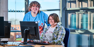 two female students on campus at the student pcs one sits at desk the other, smiling is leaning over