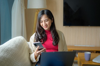 lady in a home setting sitting on sofa with laptop op lap and holding up a mobile phone