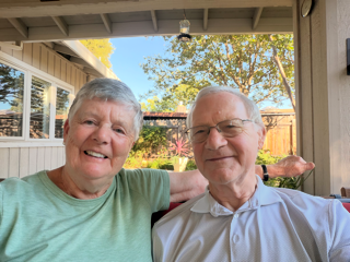 Alan and Jean sit outdoors. Jean has on a green t-shirt and puts her arm around Allan. Allan wears a white shirt and smiles for the camera