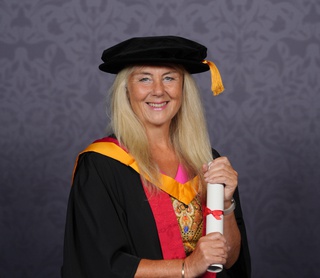 Amanda poses for a professional graduation photo dressed in graduation robes in University of Wolverhampton colours, yellow and orange. She holds a scroll between her two hands and smiles for the camera.