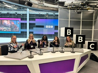 Five female BA BSL students are seated in ta a row, with microphones in front of then in the newsroom at the BBC studios at BBC Wales. They are using their hands to sign for the camera and smiling.