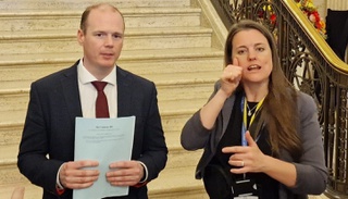 Gordon Lyons MLA stands in a suit in front of some steps, he is holding some paper and speaking to cameras. Sally Gillespie is beside him interpreting for the cameras.