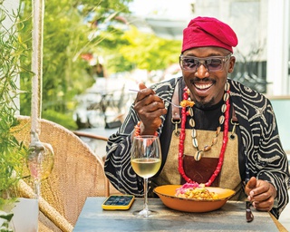Chef Eros is sitting at a table with a window behind him. He wears a red chef's hat, brown apron, dark sunglasses and a large gold chain. He has a glass of white wine to his right and bowl of food in front of him. He holds a fork of food to his mouth as he grins.