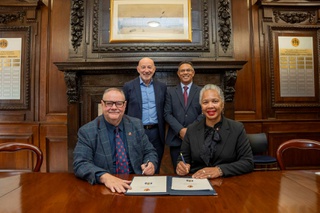 Representatives from the council and university are in an old panelled room. Two are seated with two standing behind them. On the table in front is the agreement they are signing.
