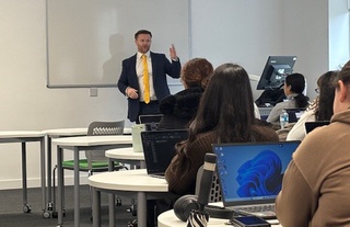 Daniel, in a suit and yellow tie, stands at the front of a classroom in front of a white board. He speaks to the students in a full class.
