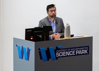 Dr Rahman, research project lead stands behind a grey podium with the University of Wolverhampton Science Park logo on the front. He is addressing an audience and wears a grey suit with a blue shirt.