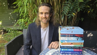 Dr Harvey is dressed in t shirt and dark suit jacket. He is outdoors seated on a chair next to a blue mosaic table. One the table are various climate fiction books stacked to make a tower.