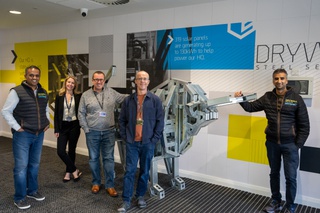 Four men and one woman stand in a room at Drywall Steel Sections Ltd for a picture. They are dressed casually and smile for the camera. Behind them is a steel framed object and the walls have the company logo and geometrics shapes in blue and yellow.