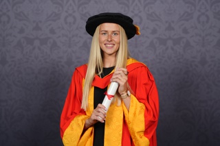 Hannah poses for a professional graduation photo dressed in graduation robes in University of Wolverhampton colours, yellow and orange. He holds a scroll between her two hands and smiles for the camera.