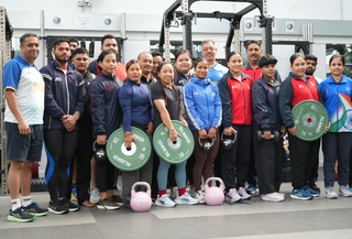 Eighteen athletes from the Indian Weightlifting Team line up for a group photo. They wear sports kit and are holding weights. Behind then are weightlifting machines.