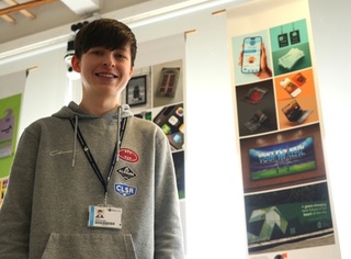 James Thomas stands in front of a display of his artwork, including the GIC logo. He smiles for the camera and wears a grey hoody