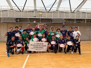Japanese male footballers gathered in a sports hall for a group photo. They wear green football shirts and hold a banner that says 'Japan-UK Inclusive Football Exchange Project.'