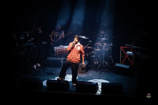 Jassi Sindhu pictured on-stage at the tribute event. He wears an orange jacket and black turban. He is in the spotlight, and the band can be seen around him in the shadows.