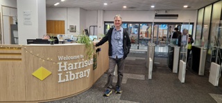 Joe stands at reception of Harrison Library at University of Wolverhampton. He smiles for the camera, wearing black jeans, blue shirt and dark blue casual jacket