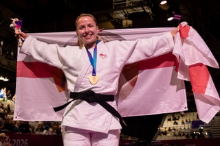 Kelly is dressed in judo whites and carries a large George Cross flag that she holds widely with two hand behind her back. She is wearing her gold medal and grins for the camera.