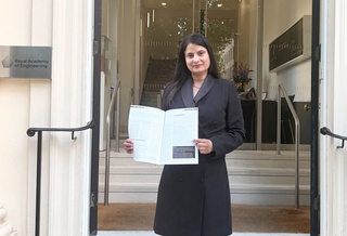 Kiran is dressed in a long black coat and stands on the steps of the Royal Academy of Engineers. She holds a magazine, within which her Green Loops innovation is featured.