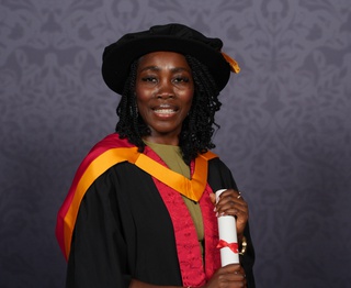 Marie-Claire wears University of Wolverhampton graduation robes in orange and yellow. She poses for a professional photo holding a scroll between her two hands and smiling for the camera.