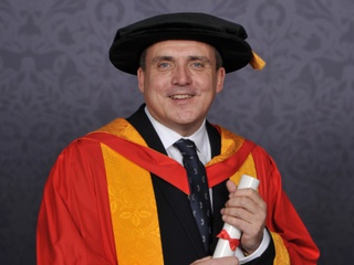 Mark Axcell poses for a professional graduation photograph dressed in University of Wolverhampton yellow and orange gown and cap. He holds a scroll in both hands