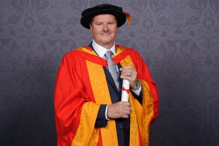 Neil poses for a professional graduation photo dressed in graduation robes in University of Wolverhampton colours, yellow and orange. He holds a scroll between his two hands and smiles for the camera.