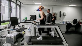 Dancer, Lauren Hewett runs on a treadmill. She has breathing monitoring apparatus over her mouth. Two men look at a computer screen whilst she runs.