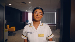A nursing student from University of Wolverhampton dressed in a white nurses uniform with a university logo and a name tag. She's standing in a room in a hospital setting.