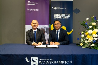 Professor Aleks Subic, Vice-Chancellor and CEO of Aston University and Ebrahim Adia, Vice-Chancellor of University of Wolverhampton sit at a table with the two University's banners behind them. They hold a pen ready to sign the agreement