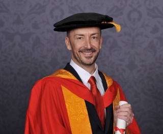 Sean poses for a professional graduation photograph in University of Wolverhampton orange and yellow gown and cap. He holds a scroll between his hands.
