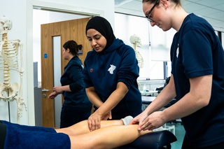 Two students wear Wolverhampton polo shirts in navy blue. They lean over a therapy table and massage someone's legs