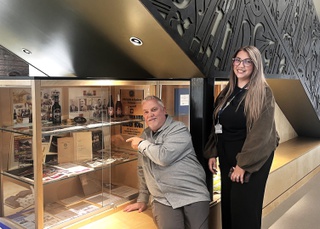 Stuart sits in front of a glass and wood cabinet. Inside are news articles, bottles and other artifacts from Mitchells & Butler Brewery. He points at cabinet, beside him stands his daughter Leigh.