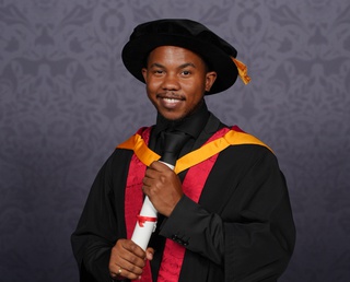 Theo poses for a professional graduation photo dressed in graduation robes in University of Wolverhampton colours, yellow and orange. He holds a scroll between his two hands and smiles for the camera.