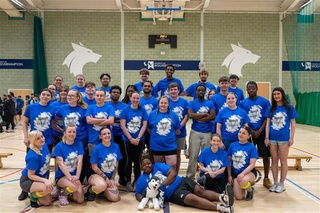 28 students from University of Wolverhampton line up in a sports hall for a photo, behind them on a green wall is a timer and university logo. They are all dressed in blue t-shirts with 'Varsity Versity' in white on the front.
