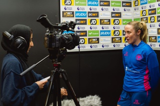 Journalism student Malaika Khan interviews Wolves Women defender, Beth Roberts. Malaika holds some notes in her right hand and is standing behind a camera wearing headphones. Beth is wearing her training kit in blue and stands in front of a backdrop of sponsor logos.
