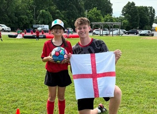 Zac is on bended knee and holds a George Cross flag. Next to him is a young girl wearing football kit and a baseball cap, she holds a football in her hands.