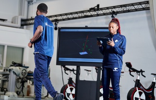 Students in sports uniforms at the sports lab, one exercising while the other looks at a tablet