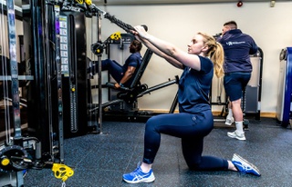 A female student using gym equipment and two male students using gym equipment behind her