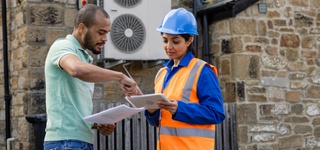 Photograph of a woman in an orange high-visibility vest and blue hardhat standing outside a house with a man pointing at a tablet in her hands.