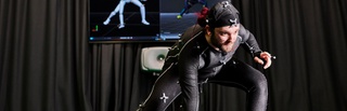 Male student using the motion capture facilities in our Screen School. They are wearing a special suit with sensors that records their movements. They have their legs apart with one hand pressed against the floor whilst the other is resting on their knee.