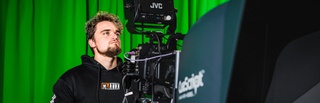 Male Film and TV Production student using a large video camera in the TV studio. You can see a green screen in the background